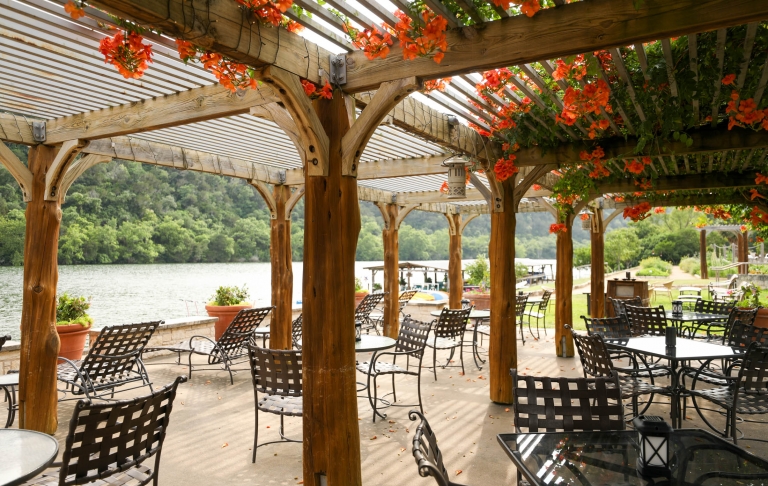 An outdoor patio where tables and chairs sit with a natural wood canopy and orange flowers peaking through its roof.