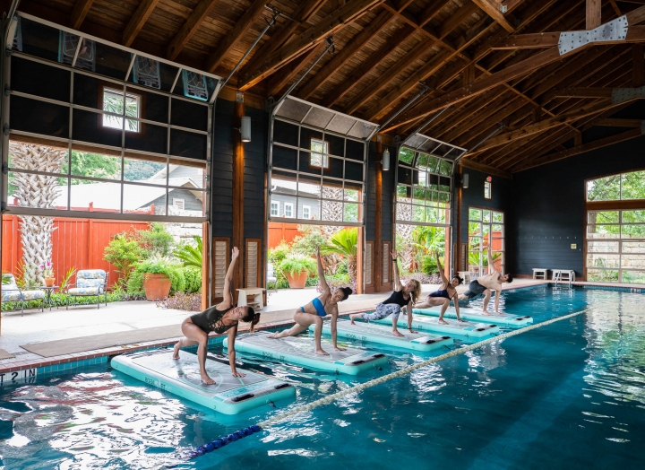 Five people in a lunge position on floating yoga mats in an indoor pool doing yoga together.