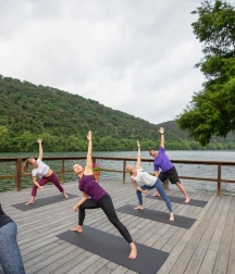 A group of people attend a yoga class on a wharf lakeside. In a lunge position, one arm is extended in the air.