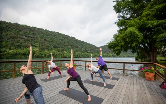A group of people attend a yoga class on a wharf lakeside. In a lunge position, one arm is extended in the air.