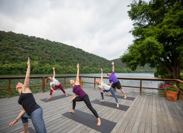 A group of people attend a yoga class on a wharf lakeside. In a lunge position, one arm is extended in the air.
