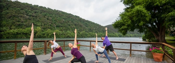 A group of people attend a yoga class on a wharf lakeside. In a lunge position, one arm is extended in the air.