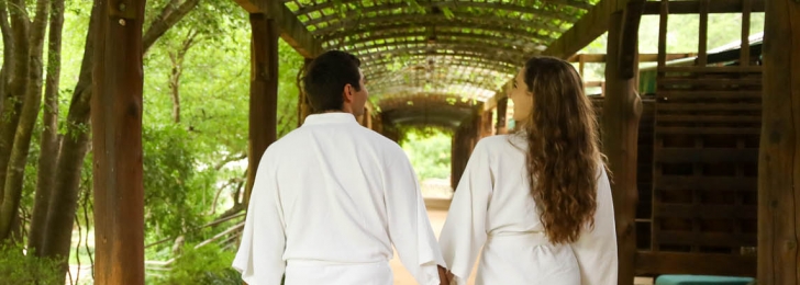 A couple holding hands walk underneath a wooden canopy surrounded by trees while holding hands.