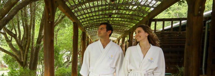 A couple walks under a wooded canopy surrounded by greenery in white Lake Austin branded robes while holding hands.