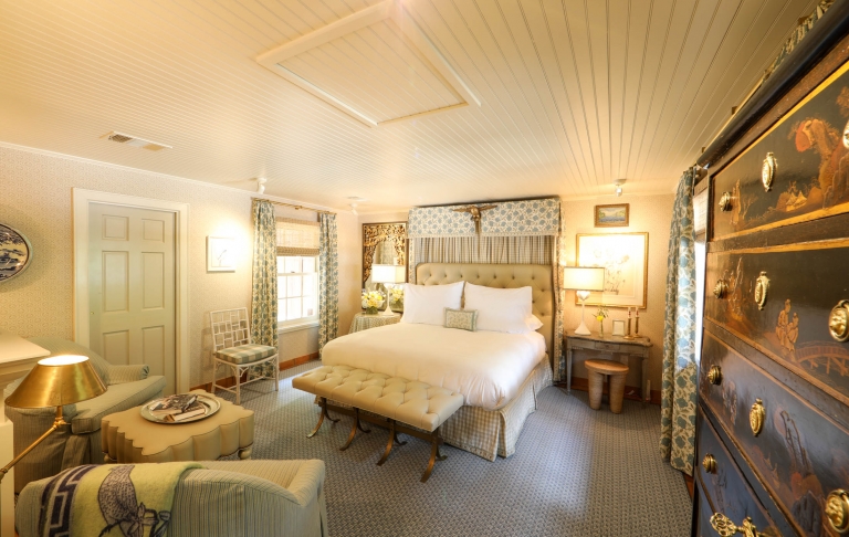 A large white bed sits in the middle of a neutral toned room with antique furniture and sage green accents.