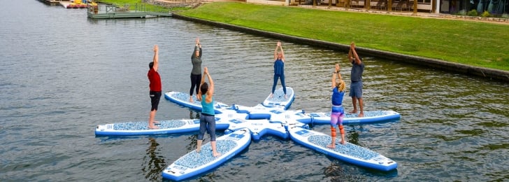 Six people perform yoga on floating boards on a lake with the resort and plenty of greenery in the background.
