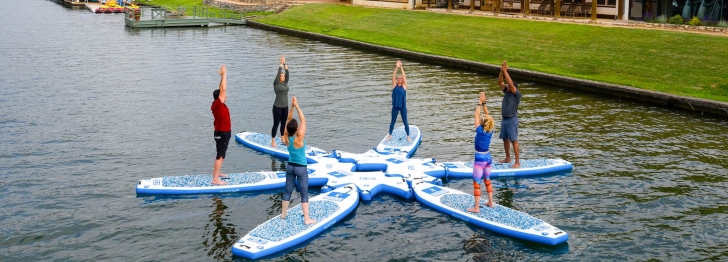 Six people perform yoga on floating boards on a lake with the resort and plenty of greenery in the background.