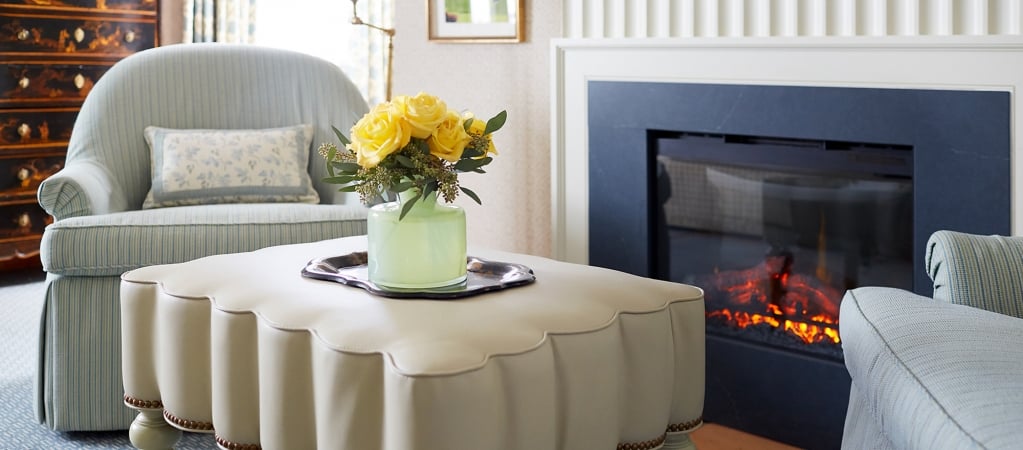 A cozy living room with a fireplace, two armchairs, a coffee table with flowers, and a warm atmosphere.