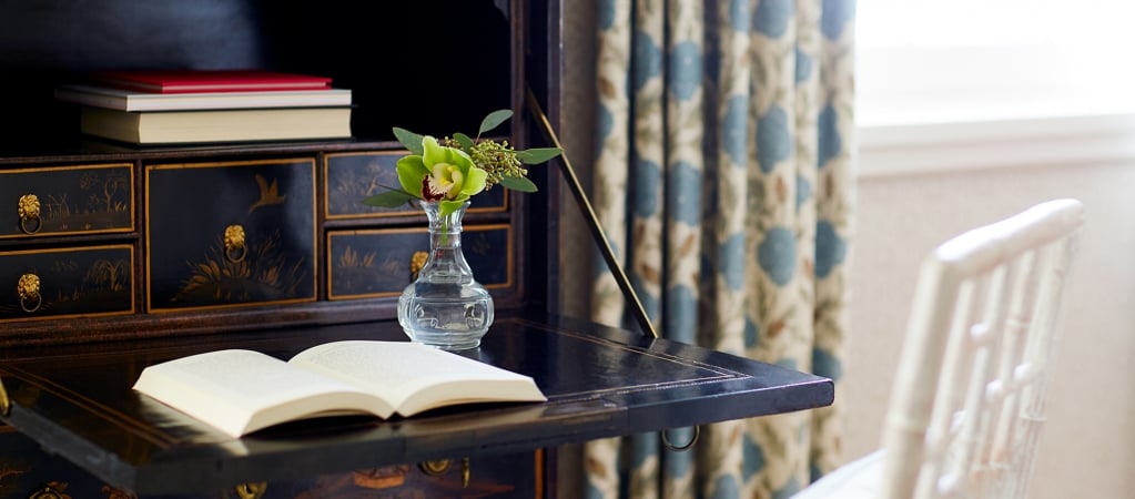 A dark wood desk with an open book, a vase with flowers, and a white chair in a room with patterned curtains.