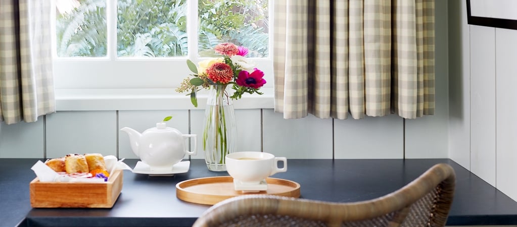 A dining area with a table set for breakfast, featuring a teapot, cup, and flowers, with a window showing greenery outside.