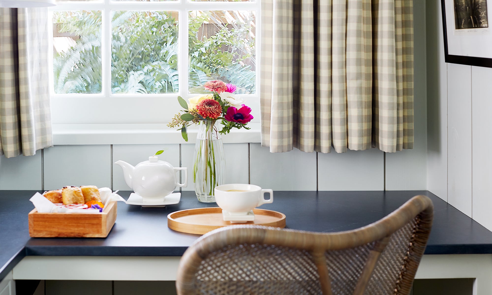 A dining area with a table set for breakfast, featuring a teapot, cup, and flowers, with a window showing greenery outside.