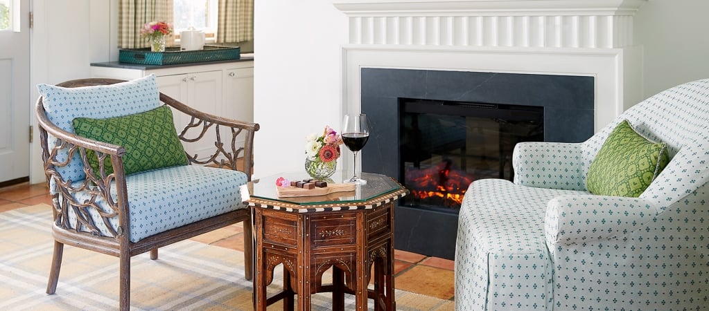 A cozy living room with a fireplace, two patterned armchairs, and a small table with a glass top.