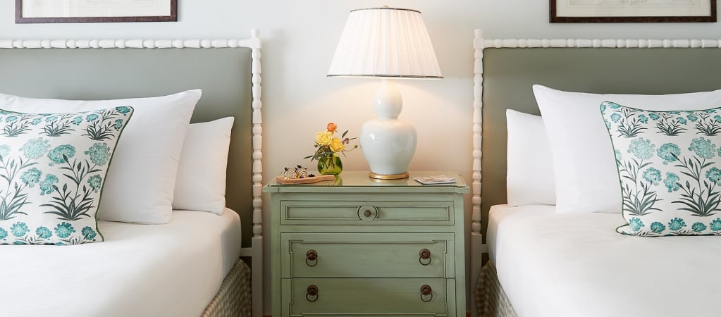 A close-up of two twin beds with white linens and green floral pillows, a green nightstand with a white lamp and flowers between them.