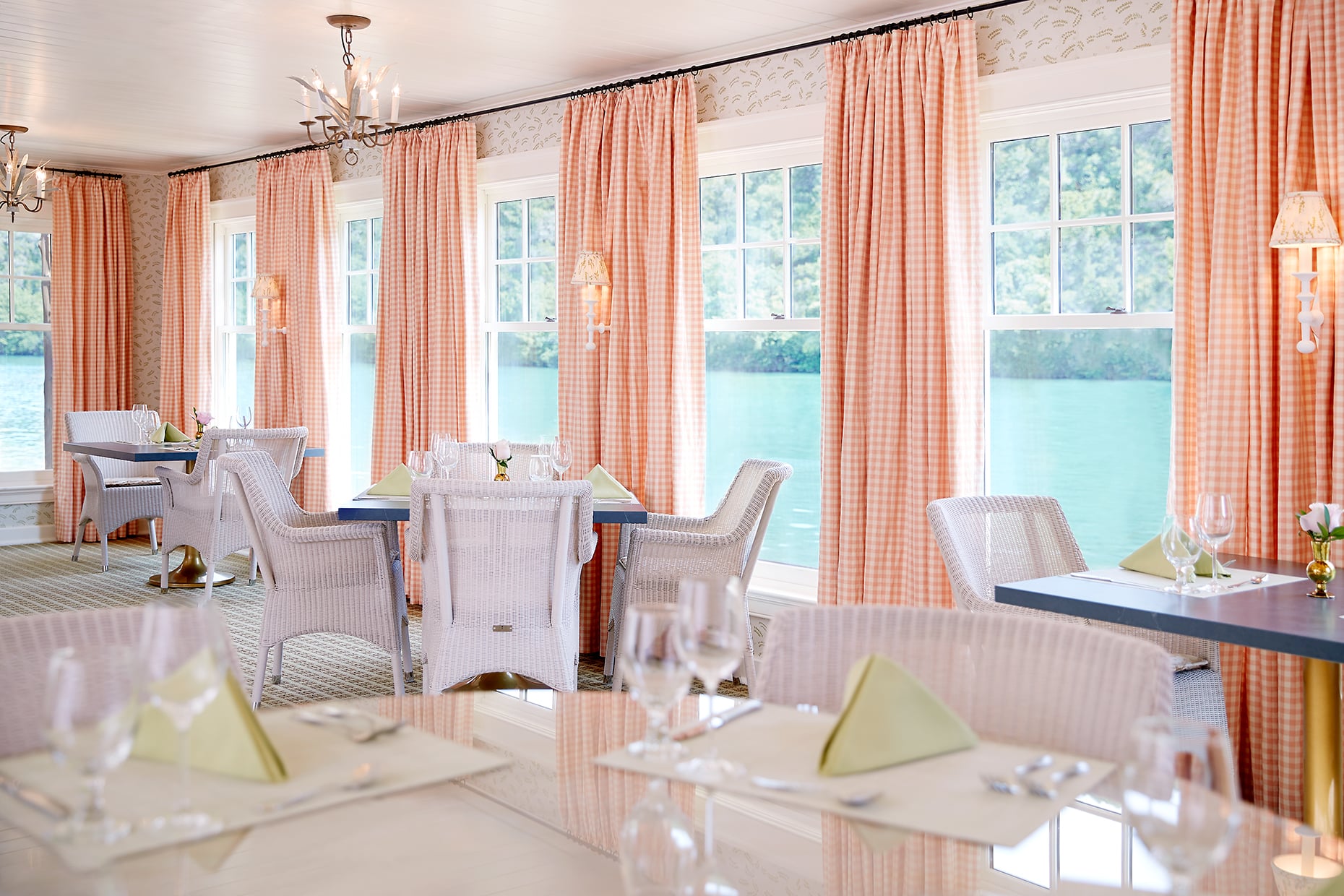 A brightly lit dining room with a lake view, featuring white wicker chairs, glass tables, and orange curtains.