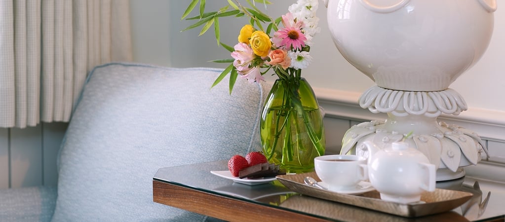 A small table with a tea set, flowers, and chocolates in a room with a blue chair.