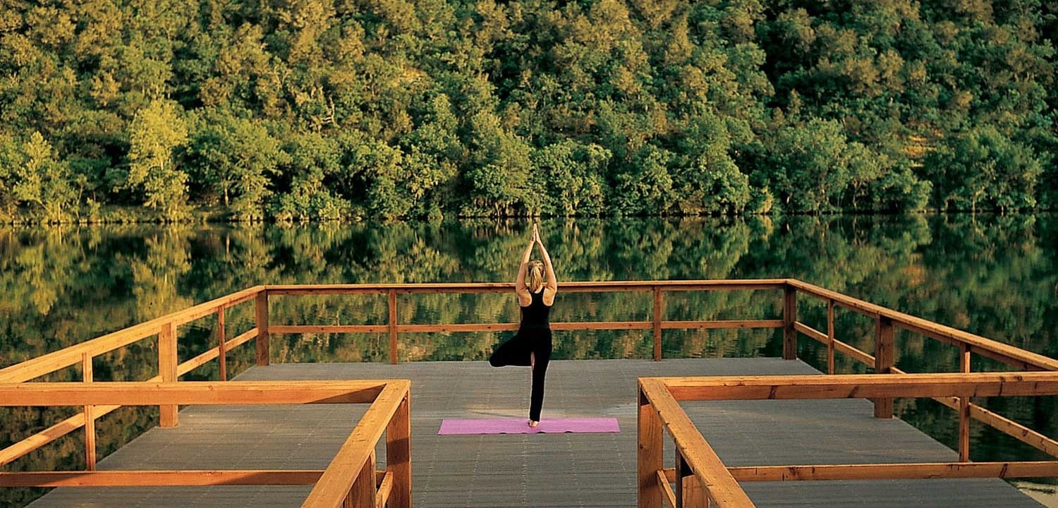A woman practices yoga on a wooden deck overlooking a lake surrounded by trees.