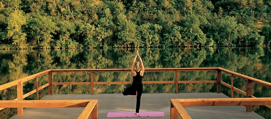 A woman practices yoga on a wooden deck overlooking a lake surrounded by trees.