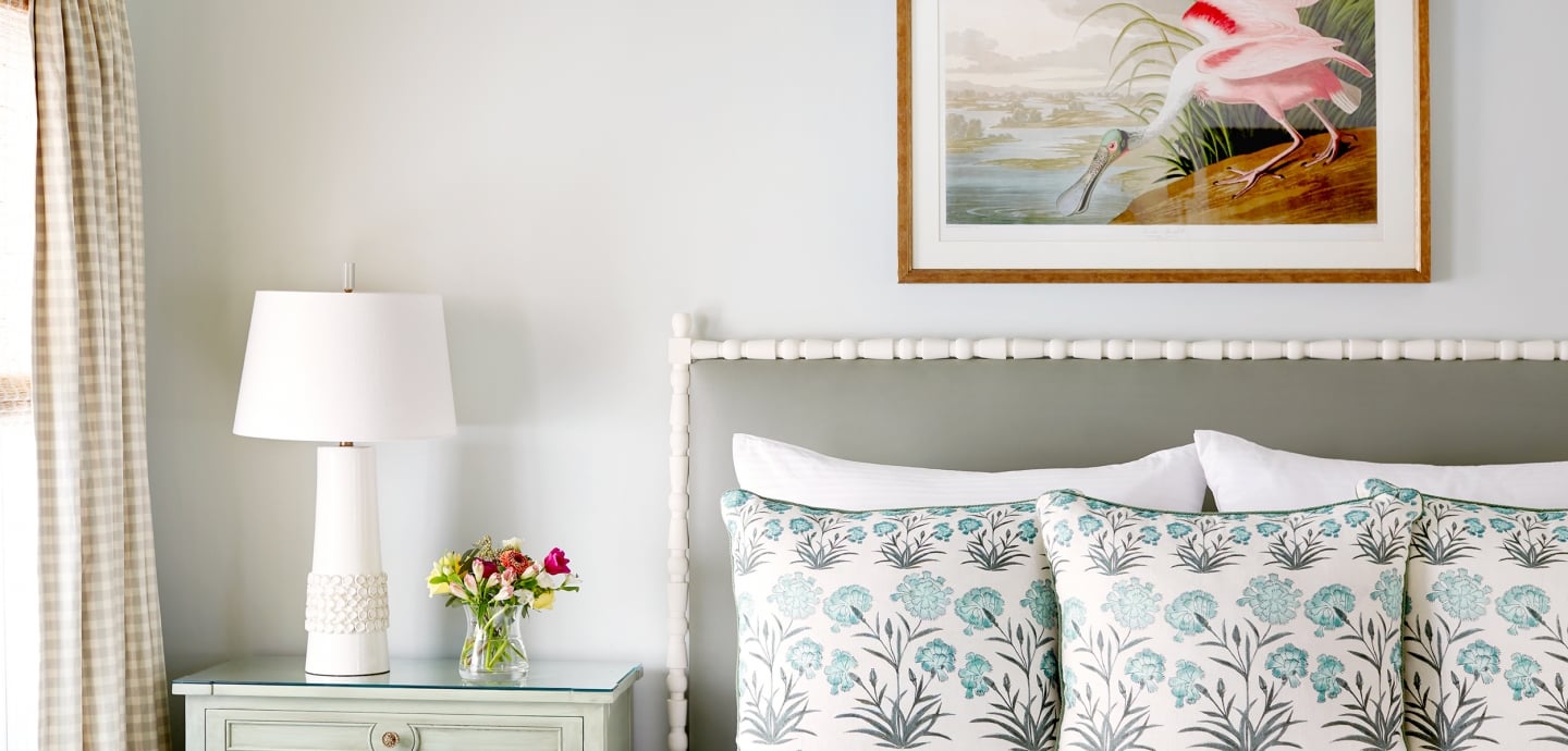 A close-up of a bed with a white and blue floral comforter, two white pillows, and a painting of a bird above the bed.
