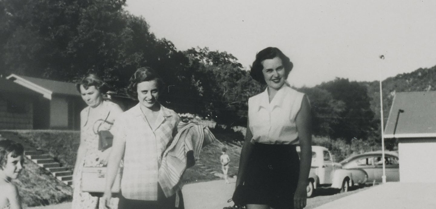 A black and white photo of three women and a child outdoors with buildings and cars in the background.