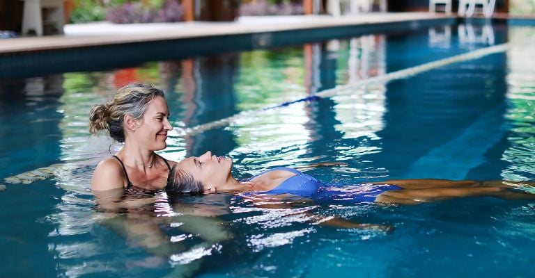 Two people are in an indoor pool, one supporting the other.
