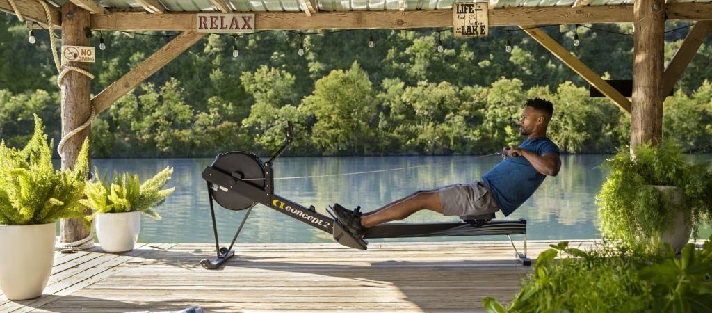A man exercises on a rowing machine on a wooden dock overlooking a lake.