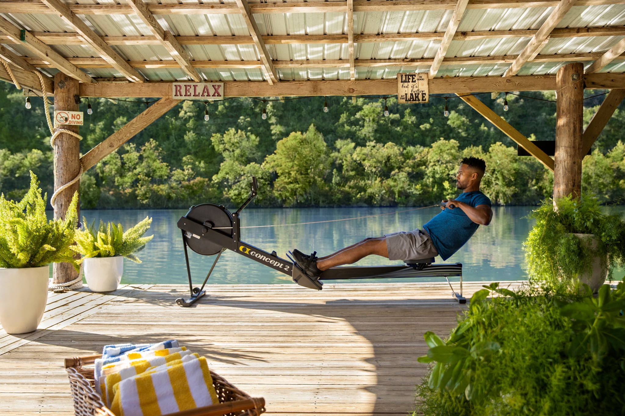 A man exercises on a rowing machine on a wooden dock overlooking a lake.