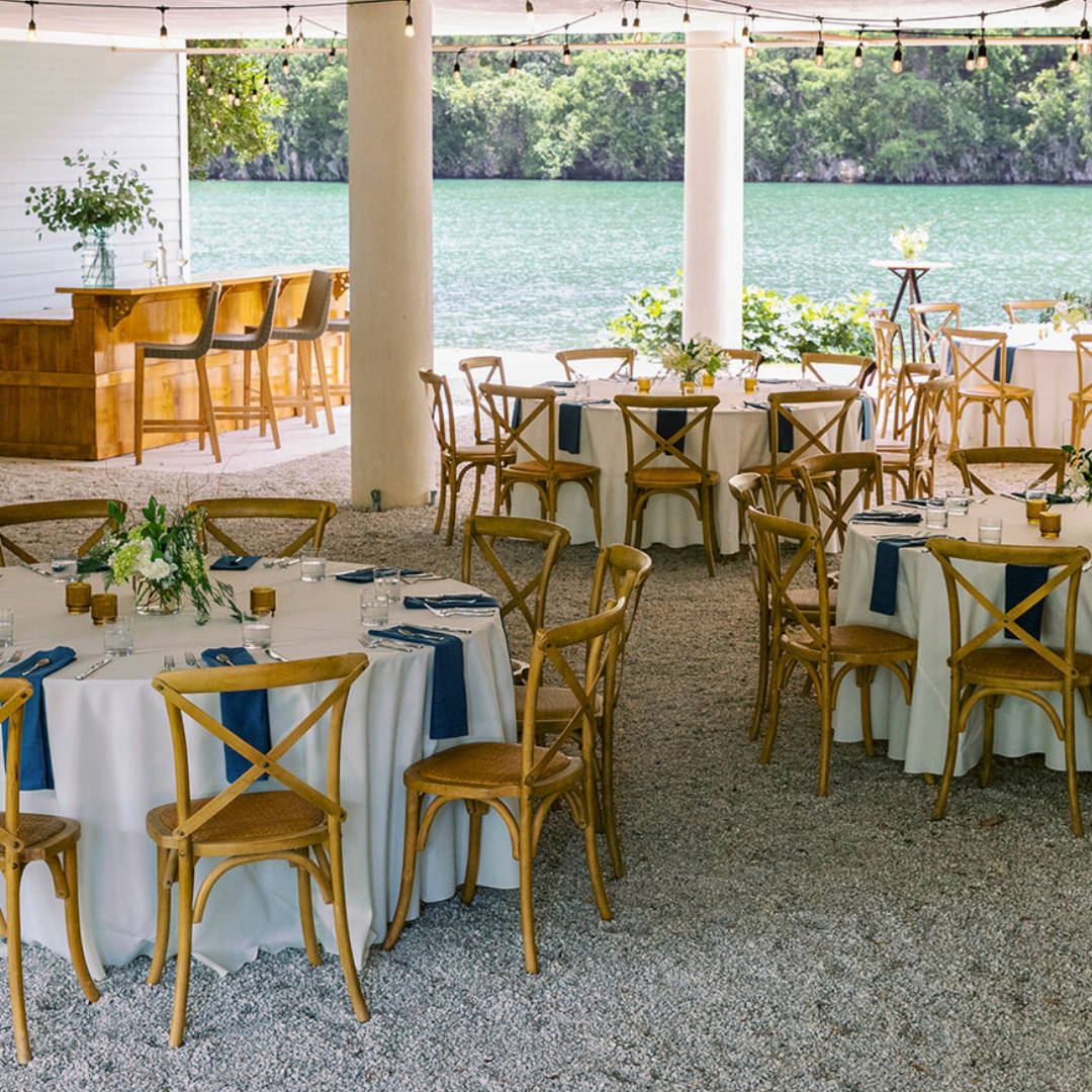 A dining area with round tables set for a meal, featuring white tablecloths, blue napkins, wooden chairs, and flower centerpieces.