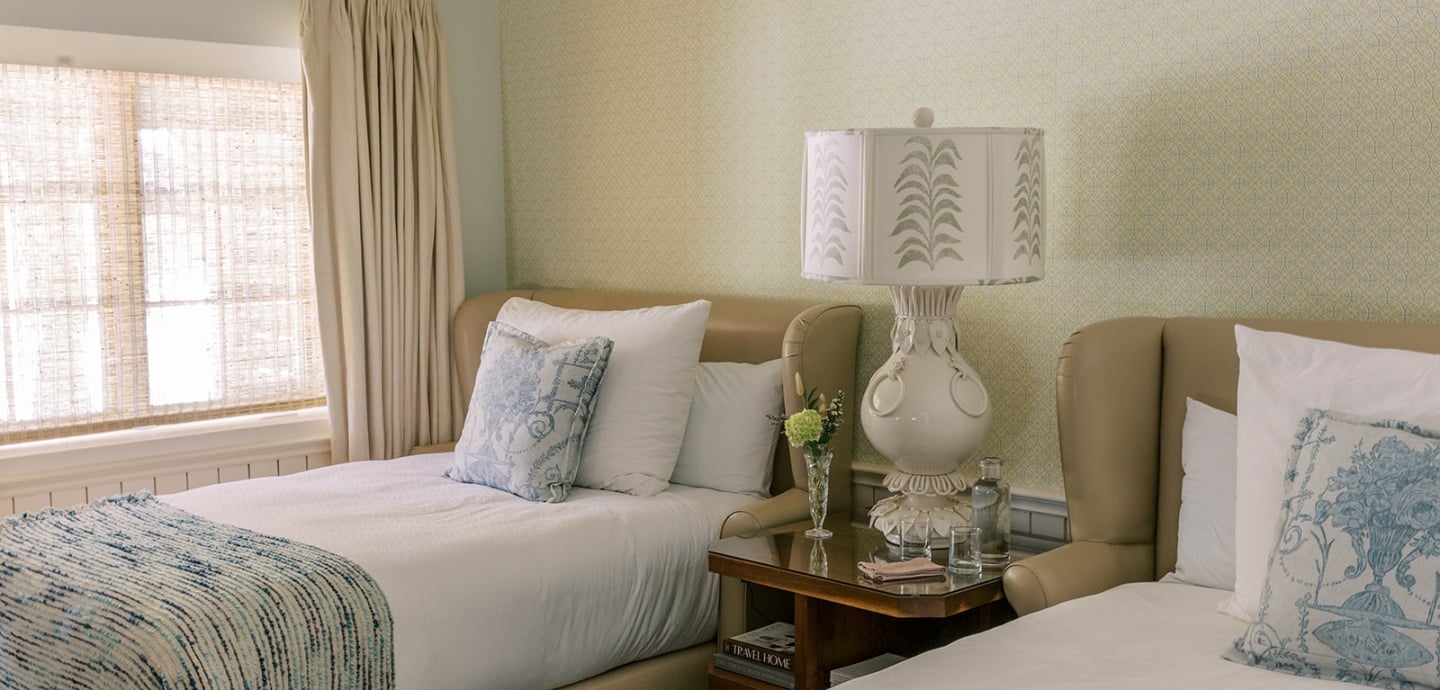 A hotel room with two twin beds, tan leather headboards, and white linens with blue accents.