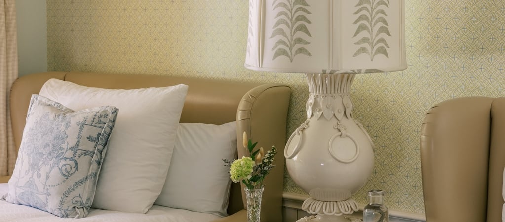 A close-up of a bed with white and blue pillows, a lamp, and flowers on a nightstand.