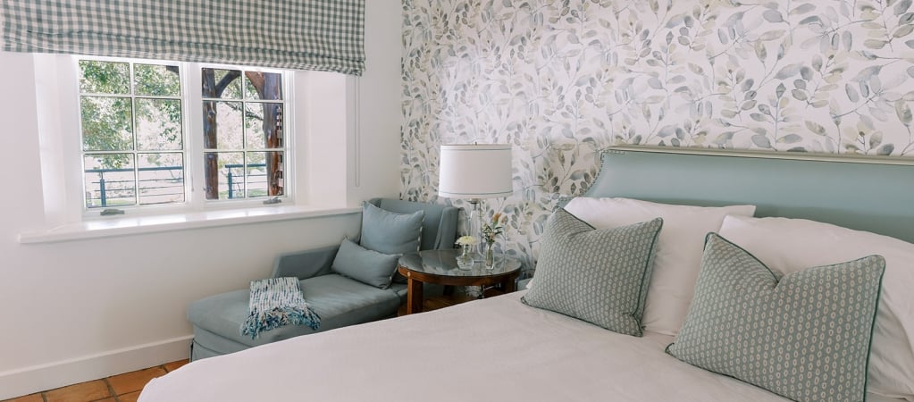 A bed with pillows and a chair in a room with wallpaper and a window.