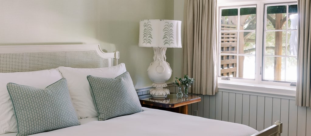 A neatly made bed with white and green pillows, a lamp, and a window with curtains.