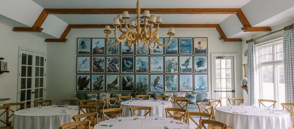A dining room with round tables, white tablecloths, and wooden chairs, featuring a large chandelier and framed pictures on the wall.