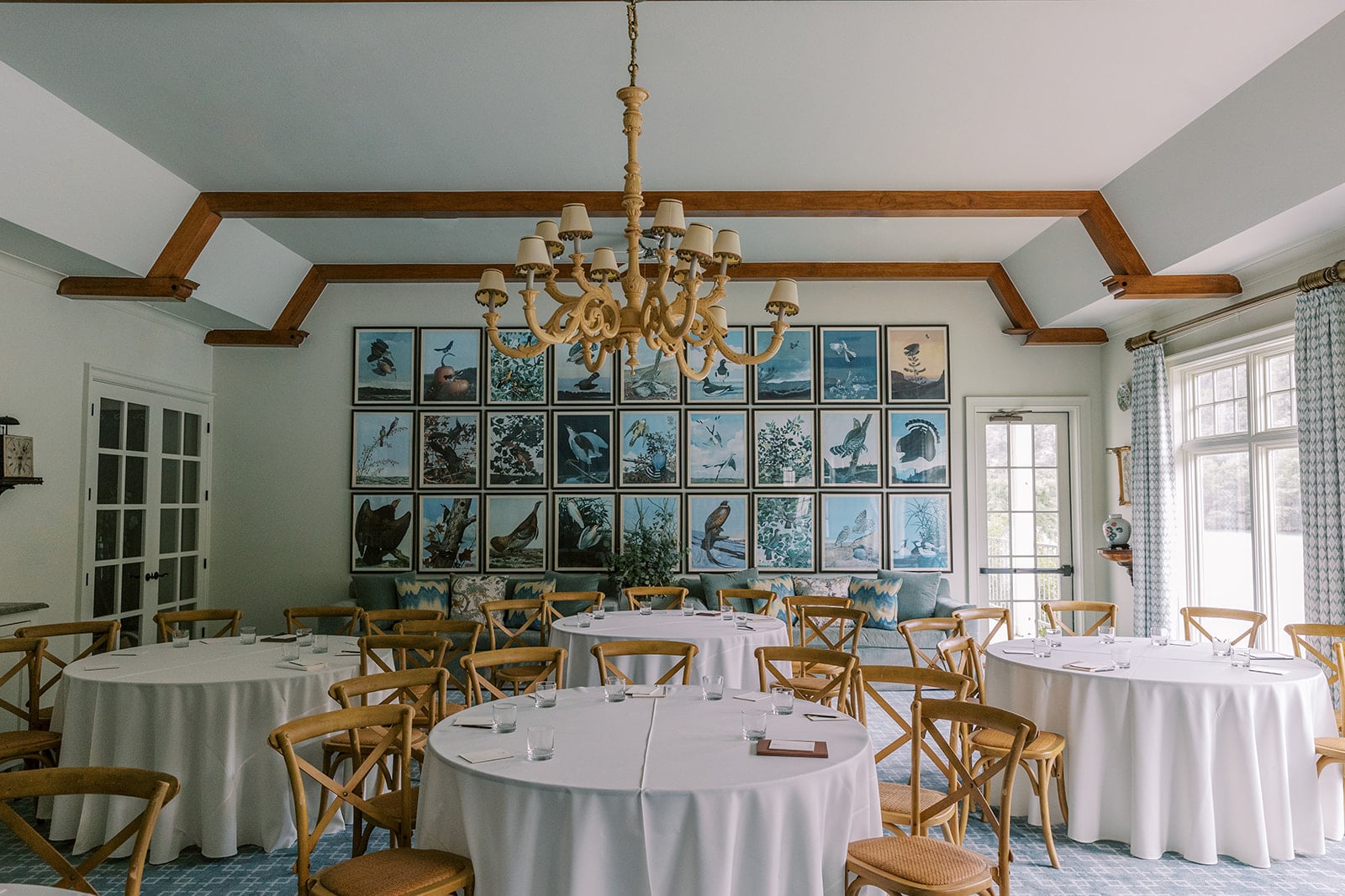 A dining room with round tables, white tablecloths, and wooden chairs, featuring a large chandelier and framed pictures on the wall.