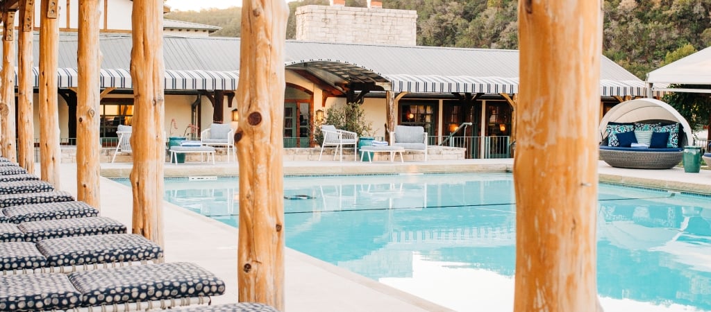 A pool area with wooden pillars, lounge chairs, and a building in the background.