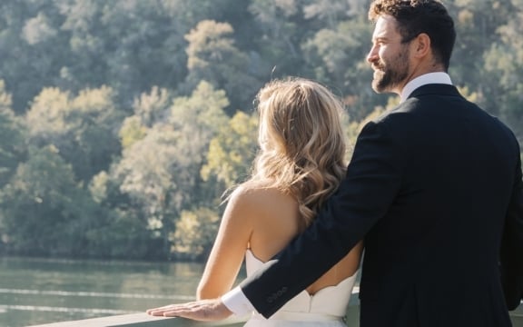 Bride and groom standing by the lakeside, facing away from the camera and looking in the same direction