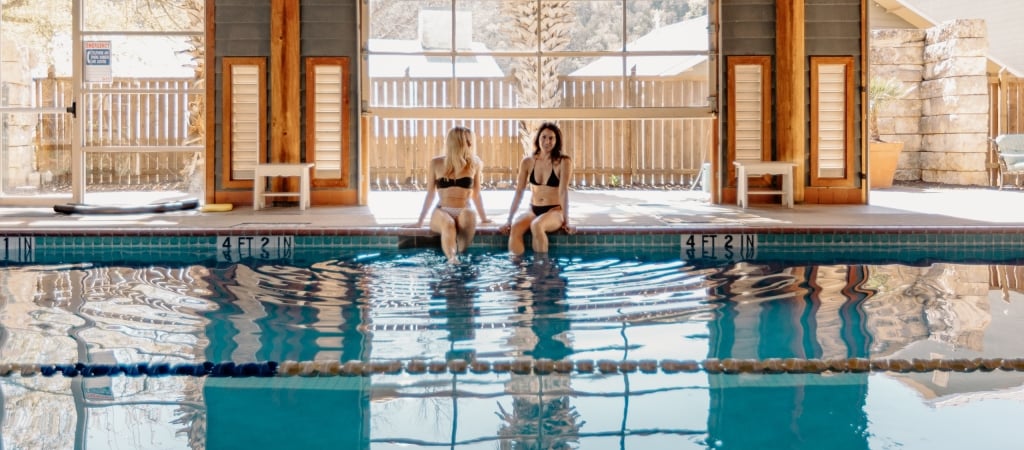 Two women in bikinis sitting at the edge of an indoor pool with large windows in the background.