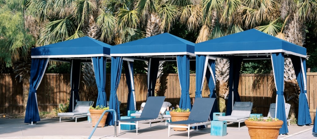 A row of blue cabanas with lounge chairs and potted plants in front of a wooden fence.